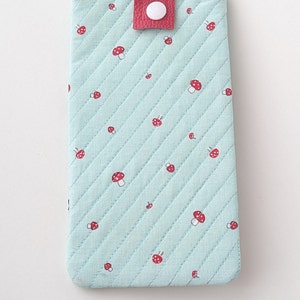 May include: A light blue quilted phone case with a red flap and a white button closure. The case is decorated with a repeating pattern of red and white mushrooms.