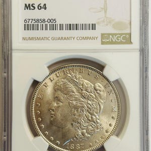 May include: A silver 1887 $1 Morgan silver dollar coin, graded MS 64, in a protective holder by Numismatic Guaranty Company (NGC). The coin shows a woman's profile and the inscription "E PLURIBUS UNUM".