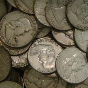 May include: A close-up of a pile of silver half dollar coins. The coins are from different years, including 1950, 1953, 1962, and 1963. The coins feature the profile of a woman with the inscription "In God We Trust" and "Liberty" on the obverse.