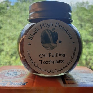 May include: A glass jar of brown oil-pulling toothpaste with a silver lid. The label reads "Black High Priestess + Essential Oregano Oil. Coconut Oil. Cloves. + Oil-Pulling Toothpaste".
