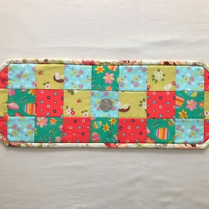 May include: A patchwork table runner with a white border. The runner is made of squares of fabric in shades of green, blue, red, and yellow. Each square has a different pattern, including flowers, eggs, and chicks. The runner is approximately 15 inches long and 6 inches wide.