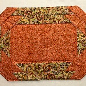 May include: Orange placemat with a paisley pattern border. The placemat has a quilted texture and a scalloped edge.