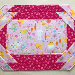 May include: Pink and white fabric placemat with a pink and white floral pattern border. The center of the placemat features a pink background with a repeating pattern of chicks, bunnies, carrots, butterflies, and eggs.