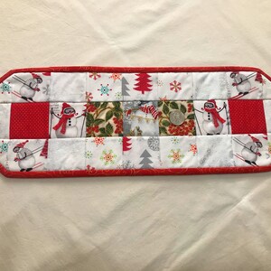 May include: A red and white quilted table runner with a winter theme. The runner features a patchwork design with snowmen, snowflakes, and Christmas trees.