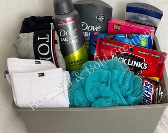 May include: A gray basket filled with various items, including a blue and gray package of underwear, white socks, a blue and green bath pouf, a blue and white bottle of body wash, a gray bottle of body wash, a red and white package of snack crackers, a blue and white package of snack crackers, a blue and white package of gum, and a blue and white package of soap.