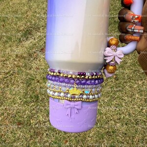 May include: A Stanley tumbler with a purple and white gradient design. The tumbler is accessorized with a stack of purple and gold beaded bracelets, a gold crown charm, and a lavender bow. The handle has a lavender bow and gold beads.