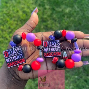 May include: A black beaded bracelet with red and purple beads. The bracelet has two charms that say "0 Days Without Sarcasm".