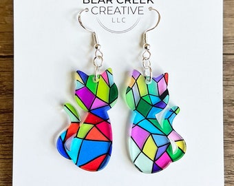 May include: A pair of cat-shaped earrings with a stained-glass design in vibrant colors. The earrings are on a white card with the Bear Creek Creative LLC logo and website address. The earrings have silver-colored hooks.