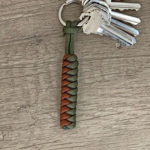 May include: A keychain featuring a braided paracord fob in olive green and brown, attached to a silver-toned metal split ring holding a cluster of five silver-colored keys. The braided cord has a cylindrical shape and a tight, uniform weave pattern.