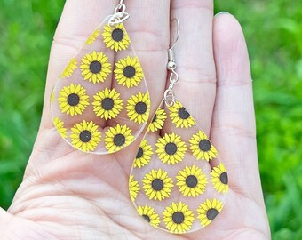 May include: A pair of teardrop-shaped earrings with a clear resin base and a sunflower pattern. The earrings have silver hooks.
