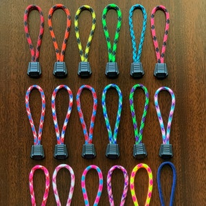 May include: A collection of twenty-four zipper pulls arranged in four rows. Each pull consists of a looped, multi-colored braided nylon cord attached to a black plastic rectangular tab with horizontal ridges for grip and texture.