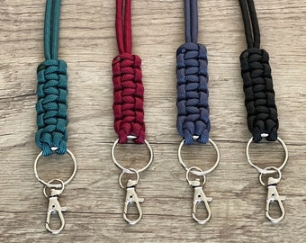 May include: Four lanyards featuring braided paracord sections in teal, burgundy, navy blue, and black. Each lanyard includes a matching cord, a silver-toned metal split ring, and a spring-loaded lobster claw clasp for attaching keys or identification.