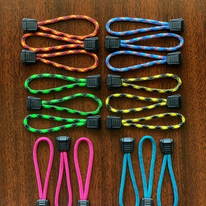 May include: A collection of fourteen zipper pulls featuring loops of braided nylon cord in various colors including orange, blue, green, yellow, pink, and teal. Each cord is secured with a black plastic rectangular toggle fastener.