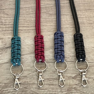 May include: Four lanyards featuring braided paracord sections in teal, burgundy, navy blue, and black. Each lanyard includes a matching cord, a silver-toned metal split ring, and a spring-loaded lobster claw clasp for attaching keys or identification.