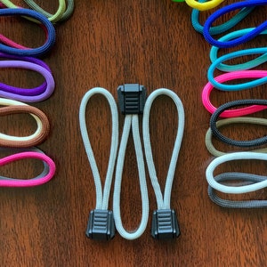 May include: A collection of colorful nylon cord zipper pulls arranged in a circular pattern. Each pull features a looped cord in various solid colors including neon green, yellow, blue, pink, purple, brown, and white, secured with a black plastic tab.