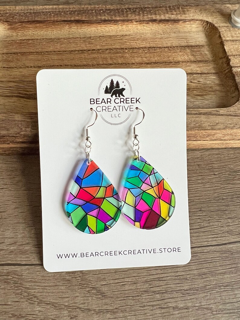 May include: A pair of teardrop-shaped earrings with a stained glass design in shades of blue, green, pink, red, and yellow. The earrings are silver and have a hook closure. The earrings are displayed on a white card with the text "Bear Creek Creative LLC" and "www.bearcreekcreative.store".