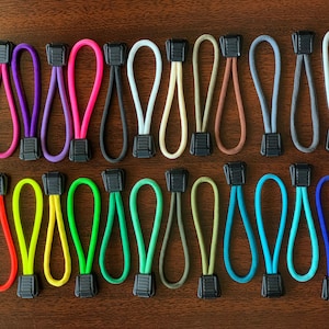 May include: A collection of twenty-six zipper pulls arranged in two rows. Each pull consists of a looped nylon cord in various solid colors, including red, pink, purple, black, white, tan, brown, gray, green, blue, and yellow, with a black plastic tab.