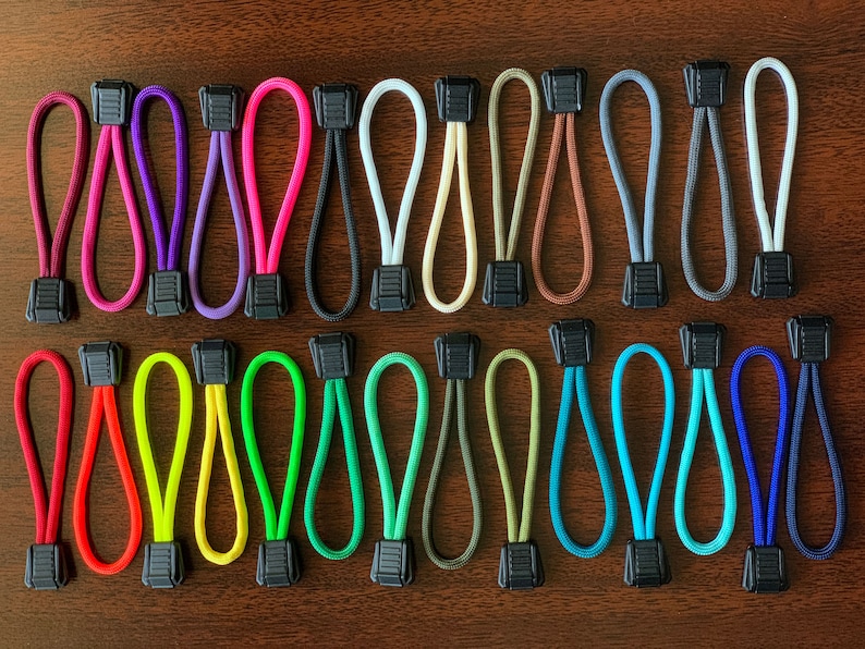 May include: A collection of twenty-six zipper pulls arranged in two rows. Each pull consists of a looped nylon cord in various solid colors, including red, pink, purple, black, white, tan, brown, gray, green, blue, and yellow, with a black plastic tab.