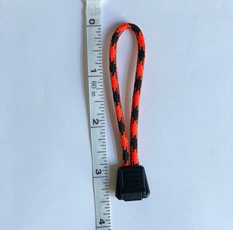 May include: A zipper pull made of orange and black braided paracord with a black plastic end cap. A white measuring tape is positioned next to the pull, showing a total length of approximately 3.25 inches from the top loop to the bottom edge.