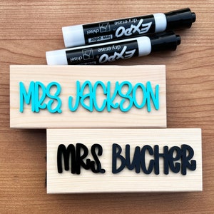 May include: Two wooden signs with the names "Mrs. Jackson" and "Mrs. Butcher" written in colorful letters. The signs are on a wooden surface with two black dry erase markers.