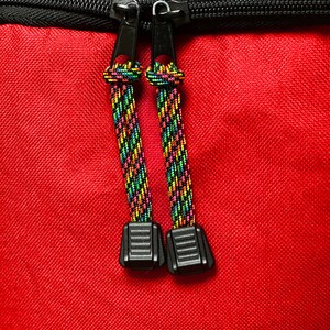 May include: A bright red fabric surface featuring a black zipper closure with two black metal pulls. Each pull is attached to a short, braided cord with a vibrant, multi-colored pattern of neon green, yellow, pink, and blue, ending in a black plastic tab.