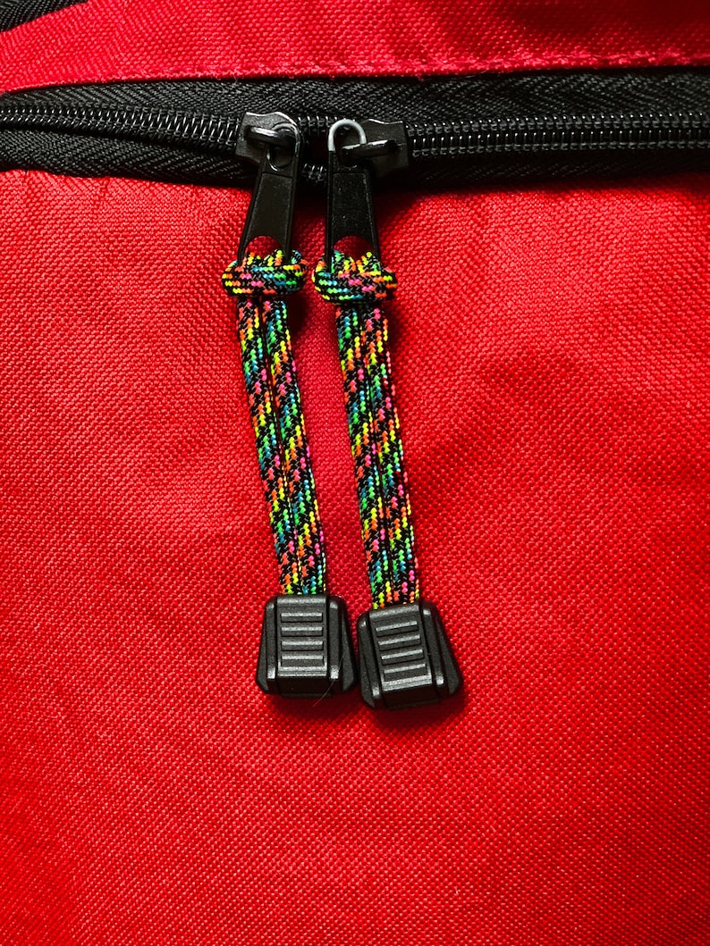 May include: A bright red fabric surface featuring a black zipper closure with two black metal pulls. Each pull is attached to a short, braided cord with a vibrant, multi-colored pattern of neon green, yellow, pink, and blue, ending in a black plastic tab.