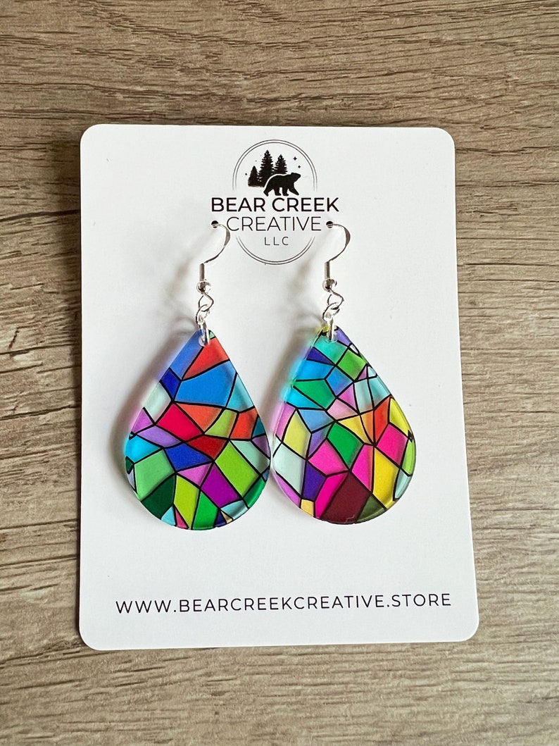 May include: A pair of teardrop-shaped earrings with a stained glass design in shades of blue, green, red, yellow, and purple. The earrings are on a white card with the text "BEAR CREEK CREATIVE LLC" and "WWW.BEARCREKKREATIVESTOR.STORE".