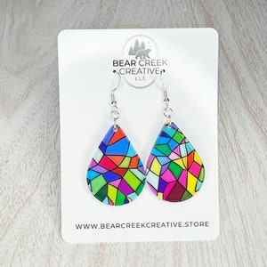 May include: A pair of teardrop-shaped earrings with a stained glass design in shades of red, blue, green, yellow, and pink. The earrings are on a white card with the text "BEAR CREEK CREATIVE LLC" and "WWW.BEARCREKKREATIVE.STORE".
