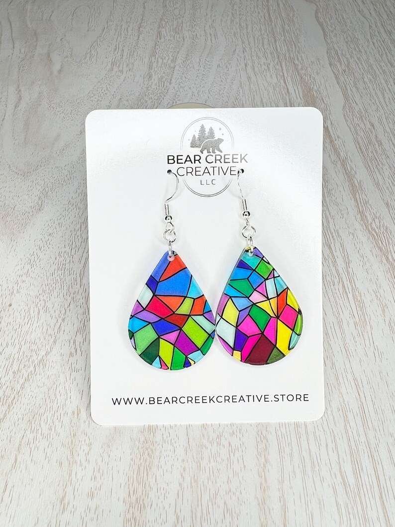 May include: A pair of teardrop-shaped earrings with a stained glass design in shades of red, blue, green, yellow, and pink. The earrings are on a white card with the text "BEAR CREEK CREATIVE LLC" and "WWW.BEARCREKKREATIVE.STORE".