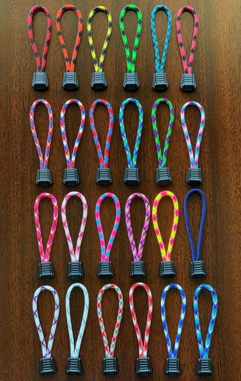 May include: A collection of twenty-four zipper pulls arranged in four rows. Each pull consists of a looped, multi-colored braided nylon cord attached to a black plastic rectangular tab with horizontal ridges for grip and texture.