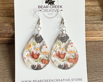 May include: Clear acrylic teardrop earrings with a repeating pattern of chickens, chicks, and foliage. The earrings are displayed on a white card with the text "Bear Creek Creative" and a website address.