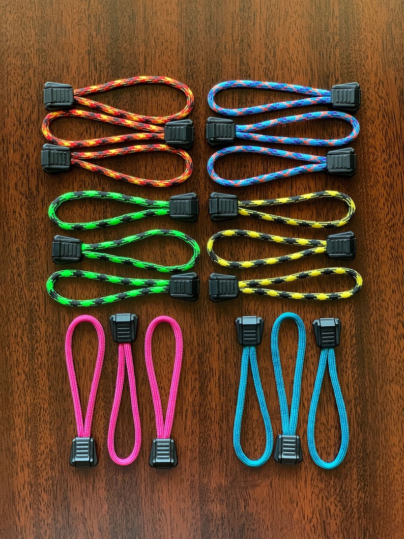 May include: A collection of fourteen zipper pulls featuring loops of braided nylon cord in various colors including orange, blue, green, yellow, pink, and teal. Each cord is secured with a black plastic rectangular toggle fastener.