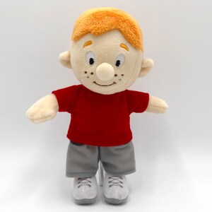 Puede incluir: Mu&ntilde;eco de peluche de un ni&ntilde;o de dibujos animados con pelo naranja, pecas y una cara sonriente. Viste una camiseta roja, pantalones cortos grises y zapatillas blancas. El mu&ntilde;eco est&aacute; dise&ntilde;ado para parecerse a un personaje amigable.