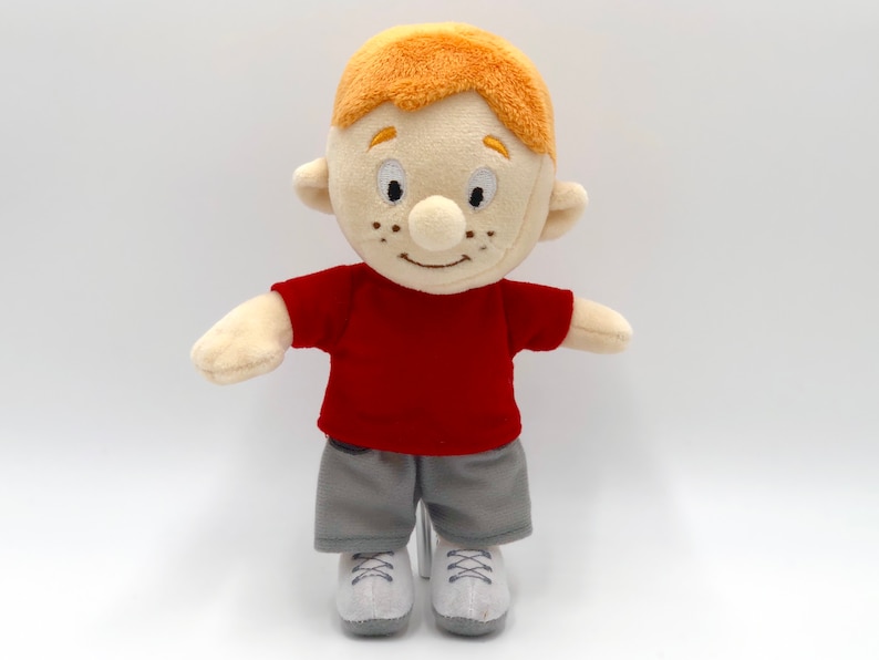 Puede incluir: Mu&ntilde;eco de peluche de un ni&ntilde;o de dibujos animados con pelo naranja, pecas y una cara sonriente. Viste una camiseta roja, pantalones cortos grises y zapatillas blancas. El mu&ntilde;eco est&aacute; dise&ntilde;ado para parecerse a un personaje amigable.
