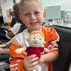 Puede incluir: Un ni&ntilde;o sonriente sostiene un mu&ntilde;eco de peluche. El mu&ntilde;eco tiene una camisa roja, pantalones grises y zapatos blancos. El ni&ntilde;o lleva una chaqueta naranja sobre una camiseta blanca. El fondo muestra una terminal de aeropuerto.