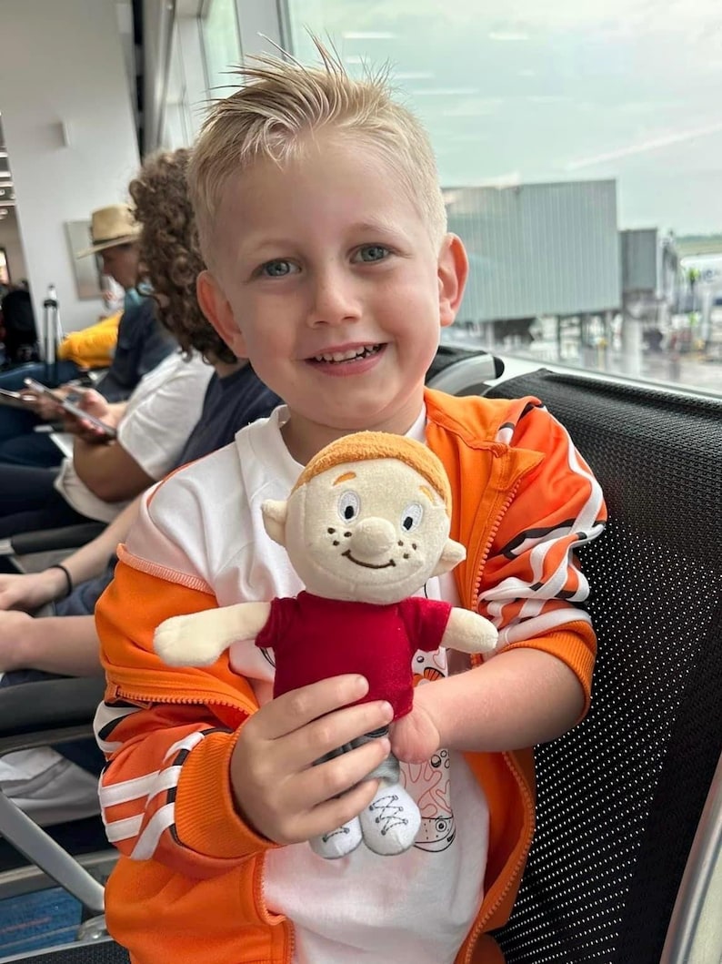 Puede incluir: Un ni&ntilde;o sonriente sostiene un mu&ntilde;eco de peluche. El mu&ntilde;eco tiene una camisa roja, pantalones grises y zapatos blancos. El ni&ntilde;o lleva una chaqueta naranja sobre una camiseta blanca. El fondo muestra una terminal de aeropuerto.