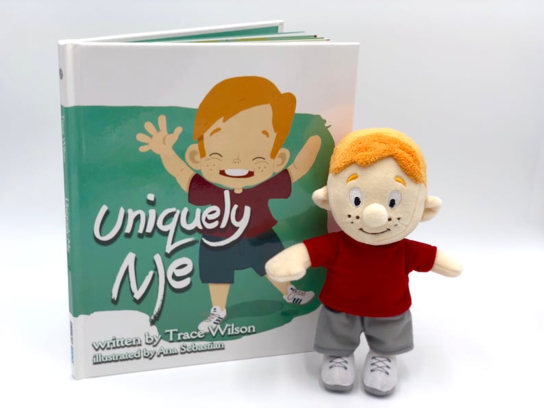 Puede incluir: Un libro infantil titulado "Uniquely Me" con un mu&ntilde;eco de peluche a juego. La portada del libro presenta una ilustraci&oacute;n de un ni&ntilde;o pelirrojo. El mu&ntilde;eco lleva una camiseta roja, pantalones cortos grises y zapatillas blancas.