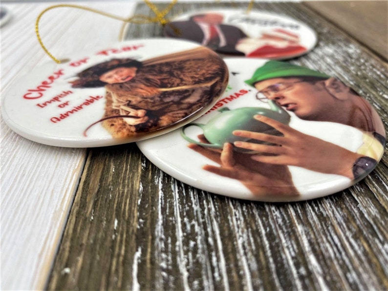 Pode incluir: Tr&ecirc;s ornamentos redondos de cer&acirc;mica com imagens e texto. O primeiro ornamento apresenta uma mulher com um casaco marrom com o texto "Cheer Up, Impish or Admirable". O segundo ornamento apresenta um homem com um chap&eacute;u verde e &oacute;culos segurando um bule verde com o texto "Merry Christmas". O terceiro ornamento apresenta um homem com terno e gravata com o texto "Merry Christmas".