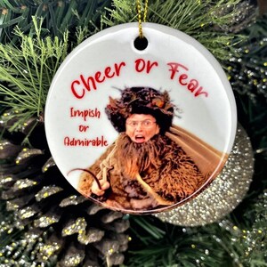Pode incluir: Enfeite de Natal de cer&acirc;mica branca com uma imagem de uma mulher com uma express&atilde;o de surpresa. O enfeite tem o texto "Cheer or Fear" e "Impish or Admirable" impresso nele.