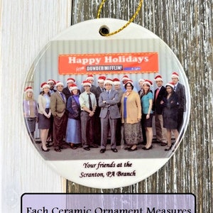 Pode incluir: Um enfeite de Natal de cer&acirc;mica branca com uma foto do elenco do programa de televis&atilde;o "The Office" usando chap&eacute;us de Papai Noel. O enfeite tem 7,6 cm de di&acirc;metro e tem o texto "Happy Holidays from Dunder Mifflin" e "Your friends at the Scranton, PA Branch".