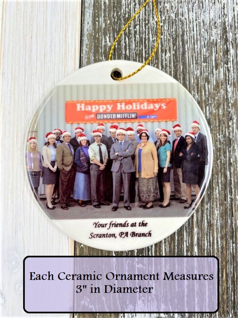 Pode incluir: Um enfeite de Natal de cer&acirc;mica branca com uma foto do elenco do programa de televis&atilde;o "The Office" usando chap&eacute;us de Papai Noel. O enfeite tem 7,6 cm de di&acirc;metro e tem o texto "Happy Holidays from Dunder Mifflin" e "Your friends at the Scranton, PA Branch".