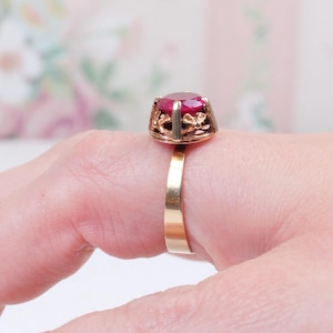 Puede incluir: Un anillo de oro con una gran gema roja redonda. La gema está engastada en un engaste calado decorativo. El anillo está en un dedo, con un fondo floral suave.