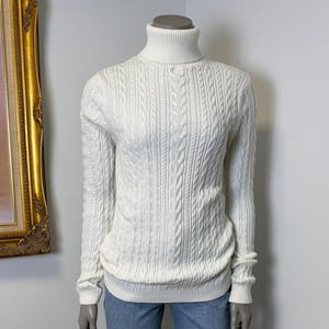 Può includere: Un maglione a collo alto color crema a trecce con maniche lunghe. Il maglione è abbinato a jeans di denim blu chiaro. Il manichino è in piedi su una base circolare nera in un angolo, con un dipinto sullo sfondo.