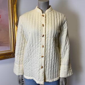 Puede incluir: Chaqueta acolchada color crema con cuello mandarín y botones de madera. La chaqueta presenta costuras verticales y mangas acampanadas. Una chaqueta de estilo vintage.