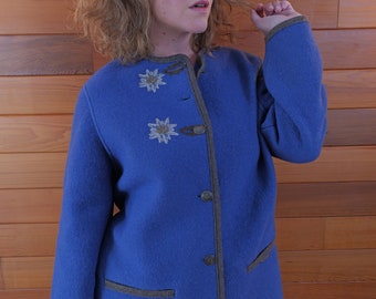 Pode incluir: Uma jaqueta de lã azul com detalhes marrons e flores brancas bordadas. A jaqueta tem gola alta e botões na frente.