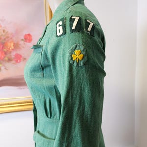 May include: Green vintage uniform jacket with the numbers "677" on the shoulder and a yellow shamrock patch. The jacket has a collared neckline, a belt, and a fitted waist. The fabric has a textured appearance.