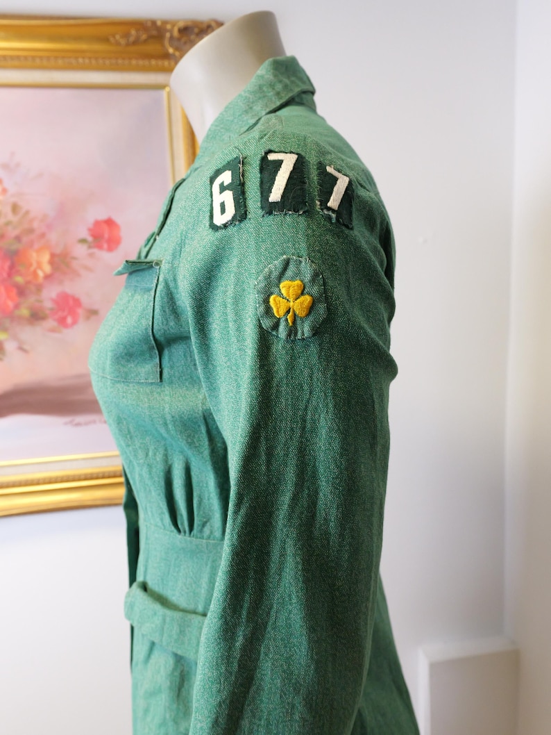 May include: Green vintage uniform jacket with the numbers "677" on the shoulder and a yellow shamrock patch. The jacket has a collared neckline, a belt, and a fitted waist. The fabric has a textured appearance.