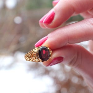 Peut inclure: Une bague en or ornée d'une pierre précieuse ovale rouge foncé. La bague présente un motif filigrané ornemental autour de la pierre et sur le corps. La bague est présentée sur un fond flou.