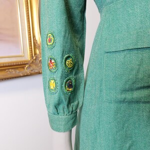 May include: A teal-green dress with a fitted waist and long sleeves. The left sleeve features several embroidered patches with colorful designs. The dress has a classic, vintage style, and a pocket is visible on the right side.