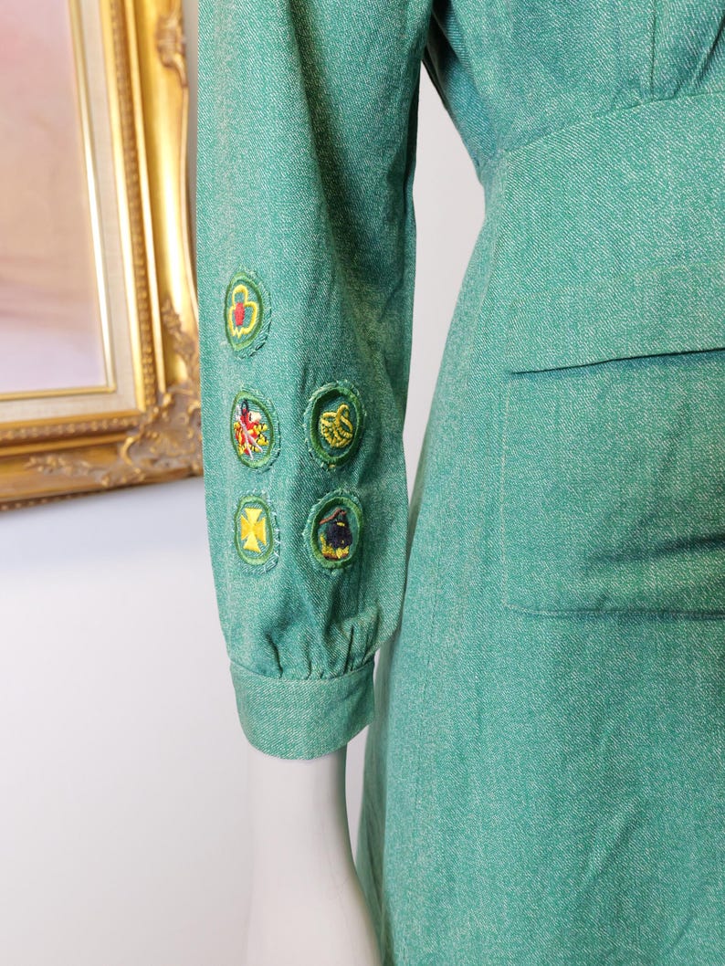 May include: A teal-green dress with a fitted waist and long sleeves. The left sleeve features several embroidered patches with colorful designs. The dress has a classic, vintage style, and a pocket is visible on the right side.
